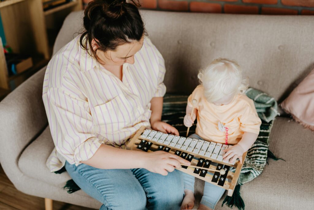 A mother and her toddler daughter playing a xylophone together indoors on a cozy sofa.